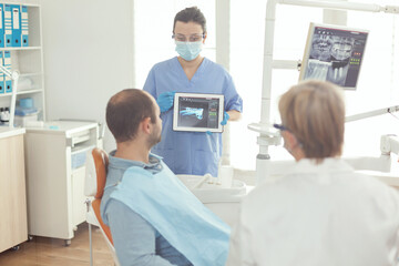 Stomatologist nurse pointing on digital screen explaining tooth x-ray to sick man sitting on dental chair in hospital stomatological office. Dentist doctor discussing toothache treatment