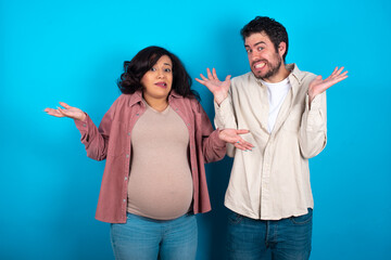 Clueless young couple expecting a baby standing against blue background shrugs shoulders with...