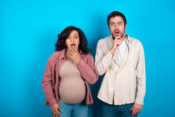 Nervous puzzled young couple expecting a baby standing against blue background opens mouth from surprise, reacts on sudden news.