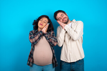 young couple expecting a baby standing against blue background leans on pressed palms closes eyes and has pleasant smile dreams about something