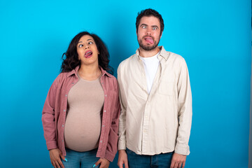 young couple expecting a baby standing against blue background showing grimace face crossing eyes and showing tongue. Being funny and crazy