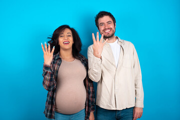 young couple expecting a baby standing against blue background smiling and looking friendly, showing number four or fourth with hand forward, counting down