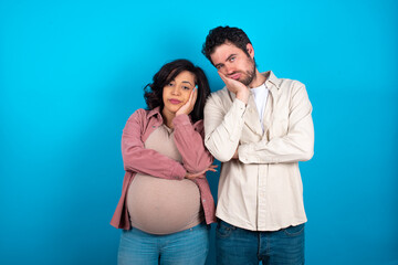 Fototapeta premium Very bored young couple expecting a baby standing against blue background holding hand on cheek while support it with another crossed hand, looking tired and sick,
