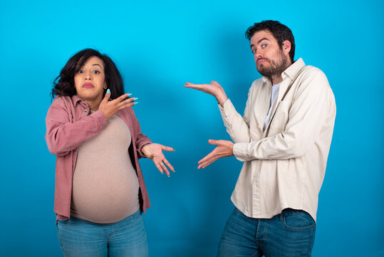 young couple expecting a baby standing against blue background pointing aside with both hands showing something strange and saying: I don't know what is this. Advertisement concept.