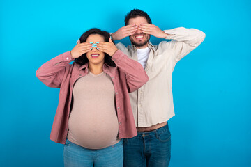 young couple expecting a baby standing against blue background covering eyes with hands smiling cheerful and funny. Blind concept.