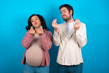 young couple expecting a baby standing against blue background doing money gesture with hands,...
