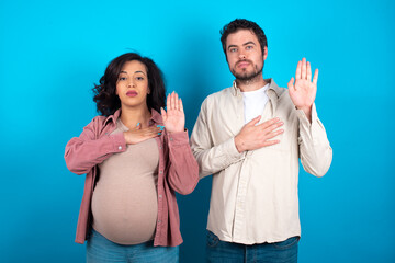 young couple expecting a baby standing against blue background Swearing with hand on chest and open palm, making a loyalty promise oath