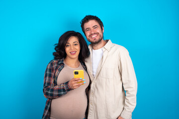 young couple expecting a baby standing against blue background Mock up copy space. Using mobile phone, typing sms message