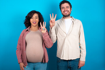 young couple expecting a baby standing against blue background showing and pointing up with fingers number three while smiling confident and happy.
