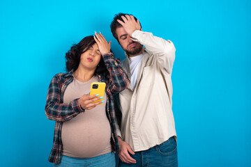 Upset depressed young couple expecting a baby standing against blue background makes face palm as forgot about something important holds mobile phone expresses sorrow and regret blames