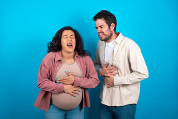 young couple expecting a baby standing against blue background with hand on stomach because nausea, painful disease feeling unwell. Ache concept.