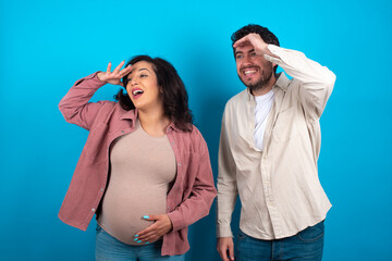 young couple expecting a baby standing against blue background very happy and smiling looking far away with hand over head. Searching concept.