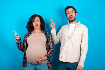 Emotive young couple expecting a baby standing against blue background keeps jaw dropped from shock demonstrates amazing promo points right on blank space demonstrates big shopping sale. Advertisement