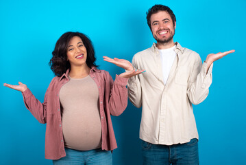 Cheerful cheery optimistic young couple expecting a baby standing against blue background holding...