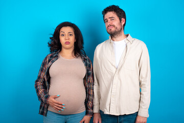 Displeased young couple expecting a baby standing against blue background frowns face feels unhappy has some problems. Negative emotions and feelings concept