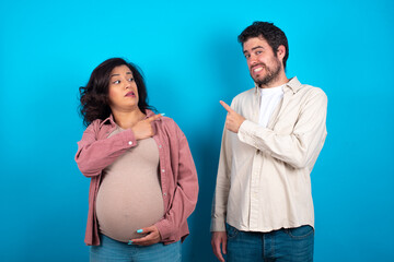 young couple expecting a baby standing against blue background Pointing aside worried and nervous...