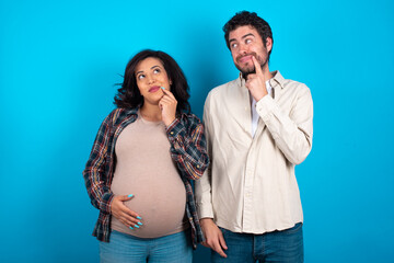 Lovely dreamy young couple expecting a baby standing against blue background keeps finger near lips looks aside copy space.