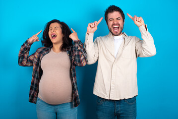 Photo of crazy young couple expecting a baby standing against blue background screaming and pointing with fingers at hair closed eyes