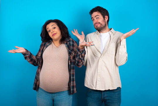 Careless Attractive Young Couple Expecting A Baby Standing Against Blue Background Shrugging Shoulders, Oops.