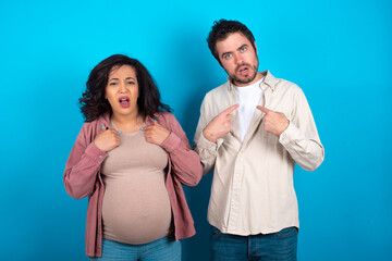 Embarrassed young couple expecting a baby standing against blue indicates at herself with puzzled expression, being shocked to be chosen to participate in competition, hesitates about something