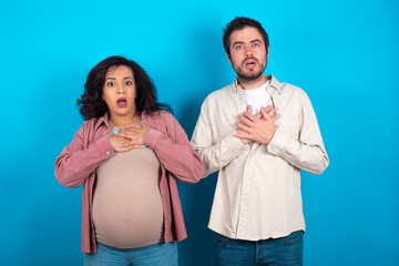 Scared young couple expecting a baby standing against blue backgr looks with frightened expression, keeps hands on chest, being puzzled to notice something strange, People, hush reaction and emotions.