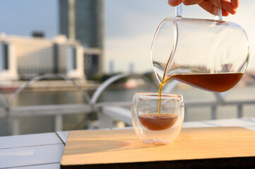 Close up pouring hot black coffee from the coffee pot into a cup on the balcony by the river. Copy space.