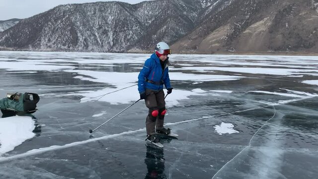 A group of tourists with sledges loaded with backpacks with equipment goes on a hike to skate on the ice of frozen Lake Baikal against the backdrop of a rocky shore