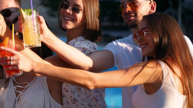 Smiling friends taking selfie with smartphone at poolside party with fresh colorful cocktails standing by swimming pool on sunny summer day. Woman taking photo at luxury villa on tropical vacation.