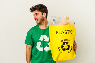 Young caucasian man recycling plastic isolated on white background looks aside smiling, cheerful...