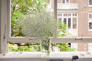 A bouquet of fresh spring white gypsophila in a vase standing on a window sill in a big bright bathroom on a sunny day. Cozy home interior decor. Blurred green tree background. Summer mood.
