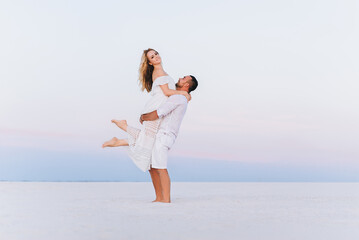 Fototapeta na wymiar A bearded man takes on his hands a blond woman on a background of white sand and bright sky. Portrait of emotional people at sunset. Love in the desert newlyweds. 