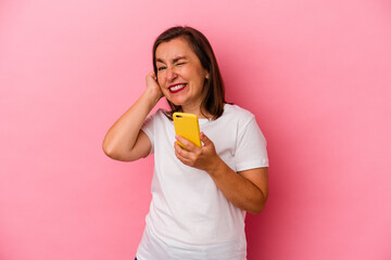Middle age caucasian woman holding mobile phone isolated on pink background covering ears with hands.