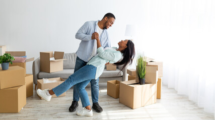 Happy loving african american couple dancing in their new house among carton boxes on moving day,...