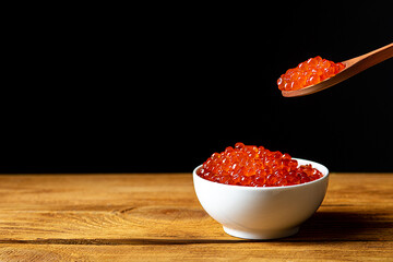 Red caviar in a wooden cup on a black background with a spoon. A large pile of bright caviar. Fresh delicious caviar. A place for advertising, logo, label, layout, mockup.