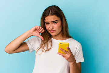 Young caucasian woman holding a mobile phone isolated on blue background showing a dislike gesture, thumbs down. Disagreement concept.