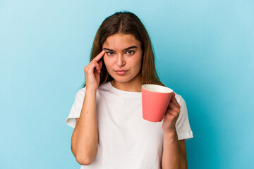 Young caucasian woman holding a mug isolated on blue background pointing temple with finger, thinking, focused on a task.