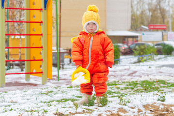 A girl in an orange jumpsuit plays with snow with a shovel on the playground