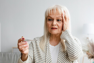 Obraz premium Scared mature woman checking body temperature looking at thermometer