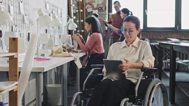 PAN slowmo of female fashion designer in wheelchair using tablet in workplace, then looking at camera while her team working on new garments