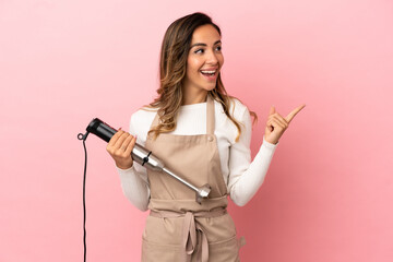 Young woman using hand blender over isolated pink background intending to realizes the solution while lifting a finger up