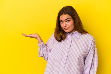 Young caucasian woman isolated on yellow background showing a copy space on a palm and holding another hand on waist.