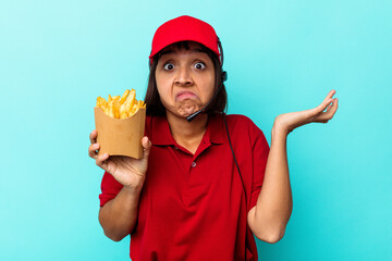 Young mixed race woman fast food restaurant worker holding fries isolated on blue background shrugs...
