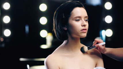 makeup artist applying powder on clavicles of pretty model with bare shoulders