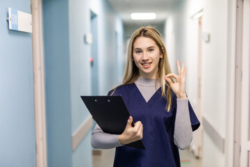 Cheerful beautiful young woman doctor with clipboard showing ok sign in hospital