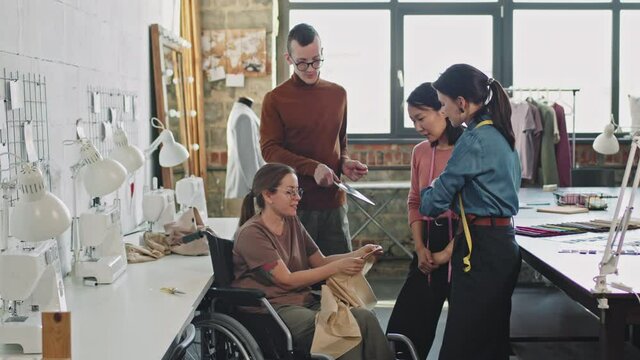 Slowmo medium shot of cheerful female seamstress in wheelchair smiling and showing garment to team of fashion designers in studio