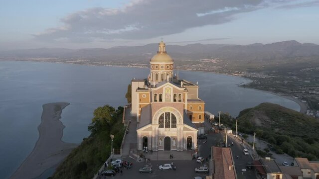 Aerial still shot of Sanctuary of Tindari baroque facade from high angle view at sunset, iconic Sicily travel destinations in Italy. Christian baroque cathedral by the coast