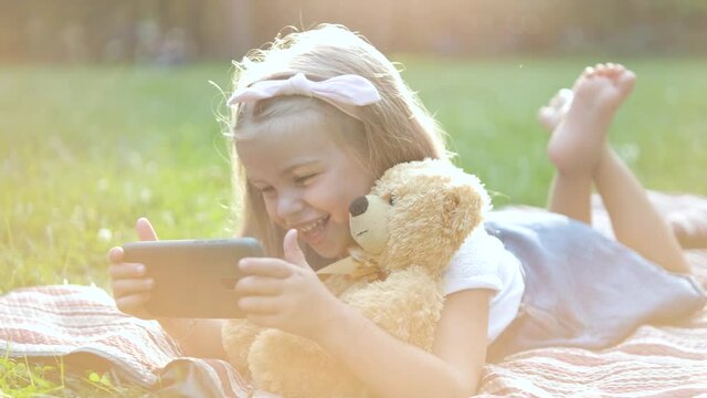 Happy little child girl looking in her mobile phone with her favorite teddy bear toy outdoors in summer park.