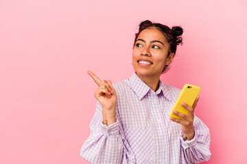 Young latin woman holding a mobile phone isolated on pink background smiling and pointing aside, showing something at blank space.
