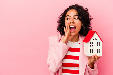 Young latin woman holding a toy house isolated on pink background is saying a secret hot braking news and looking aside - obrazy, fototapety, plakaty