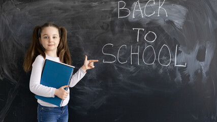 A Caucasian girl schoolgirl stands against the background of a chalk board. The words are written back to school. holds several large notebooks in his hands points to the text with his index finger.